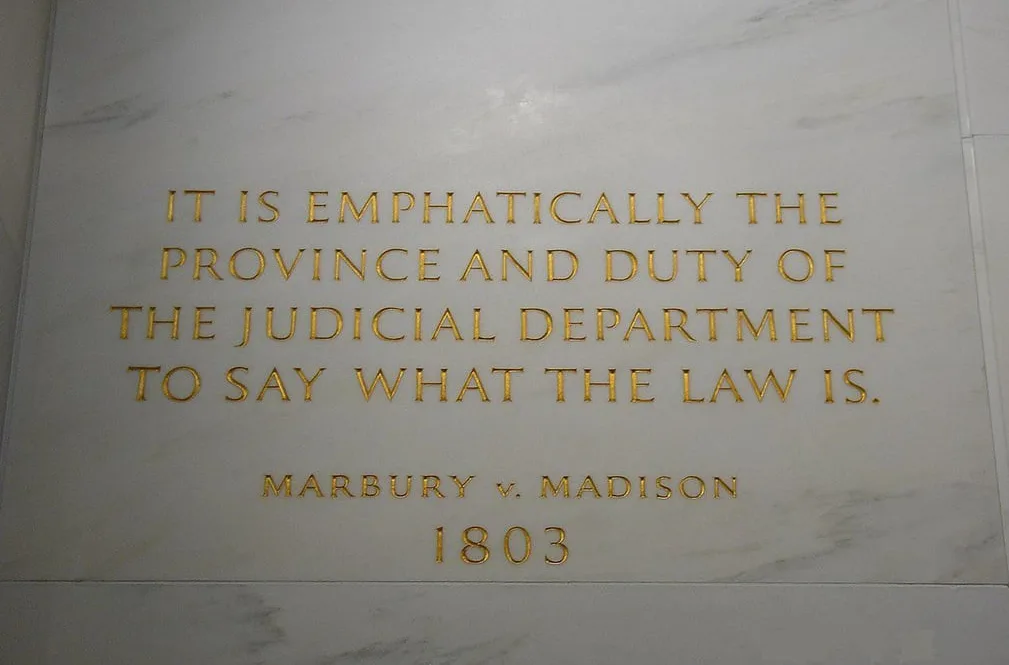 A close-up photograph of a white marble plaque with gold lettering that reads: 'IT IS EMPHATICALLY THE PROVINCE AND DUTY OF…