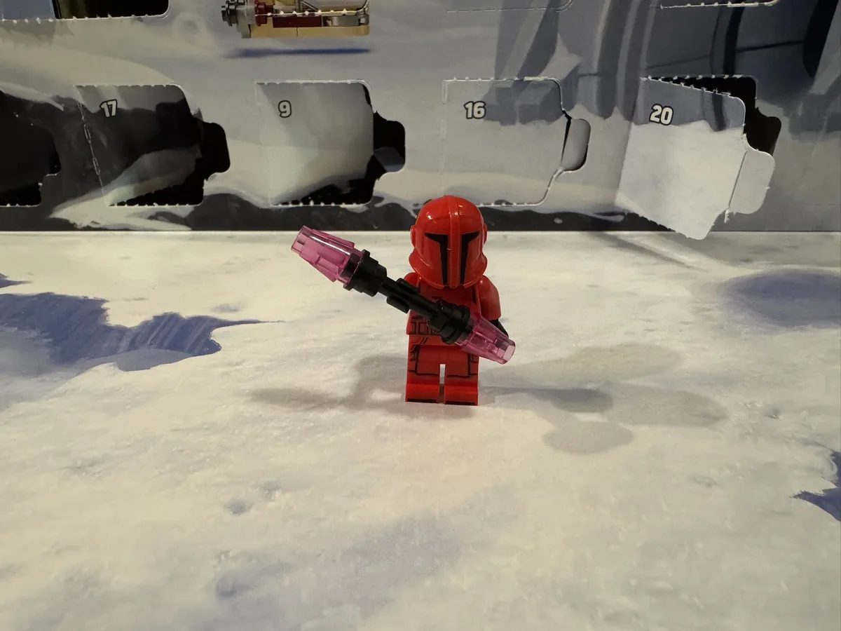 A red Lego Mandalorian minifigure stands on a snowy surface holding a pink translucent blaster, facing away from the camera…