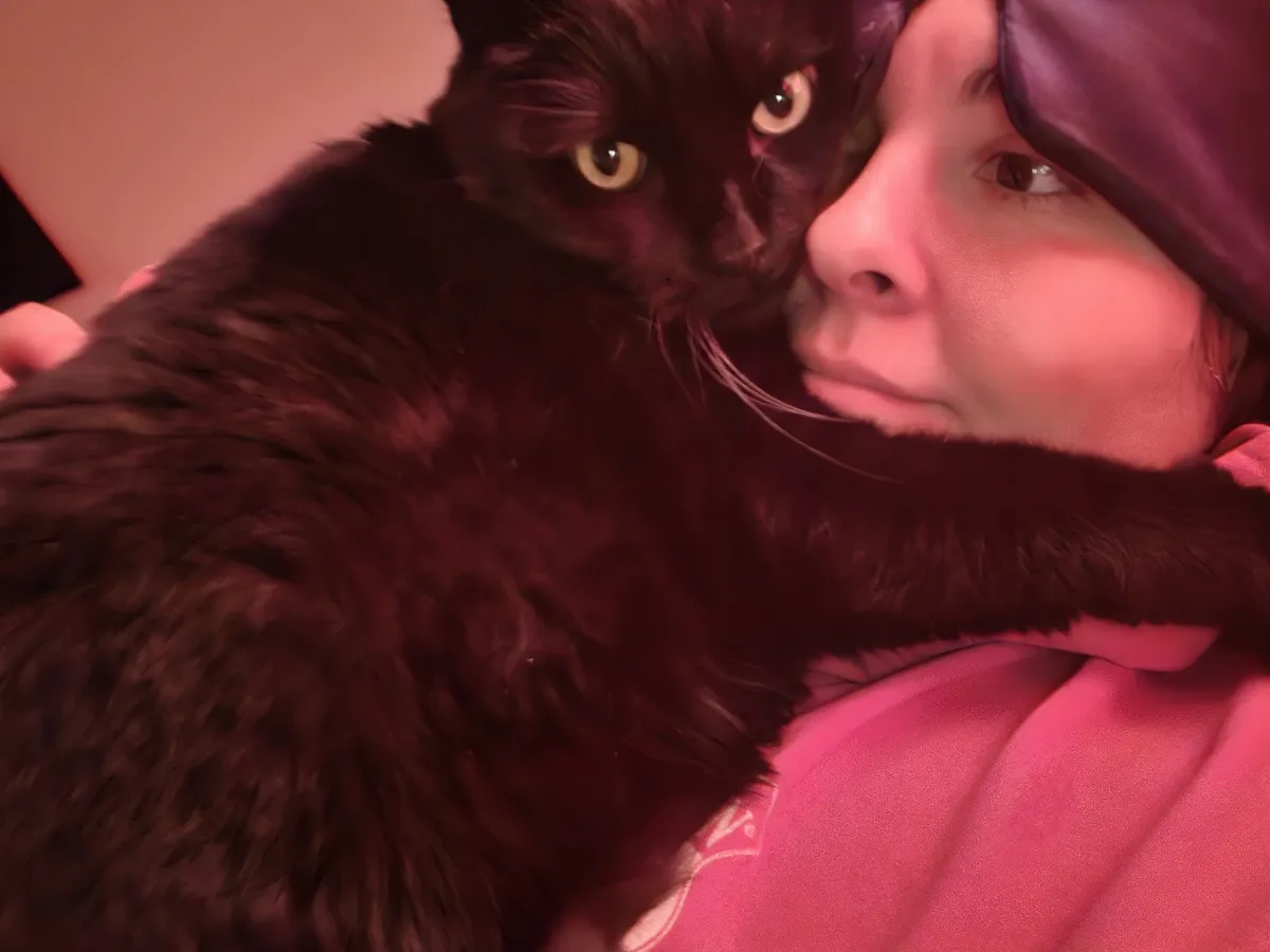 A person wearing a purple sleep mask and a pink hoodie is cuddled closely by a fluffy black cat. The cat's head is pressed…