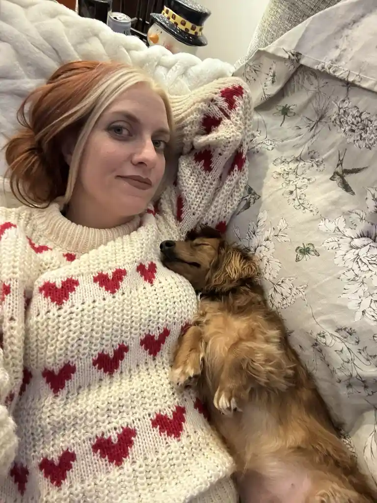A woman with red and blonde hair lies on a bed wearing a white knit sweater with red hearts, looking at the camera with a…