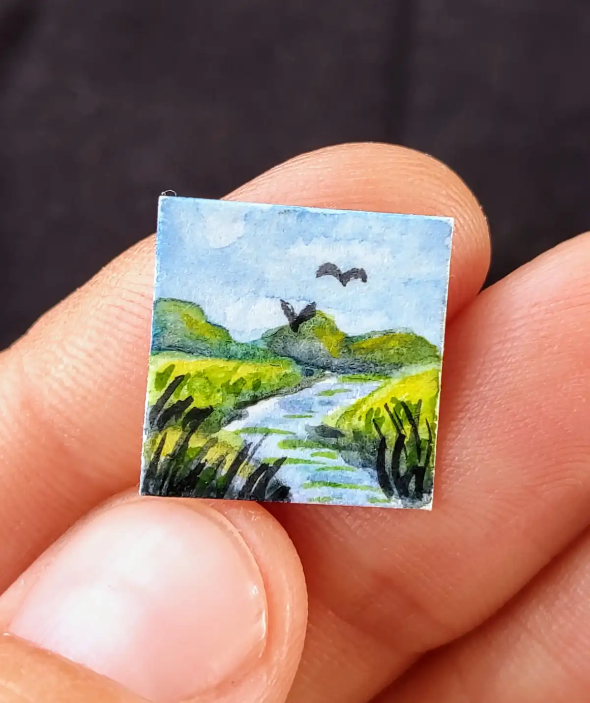 A hand holds a tiny square watercolor painting depicting a serene landscape with green hills, a winding river, and two black…