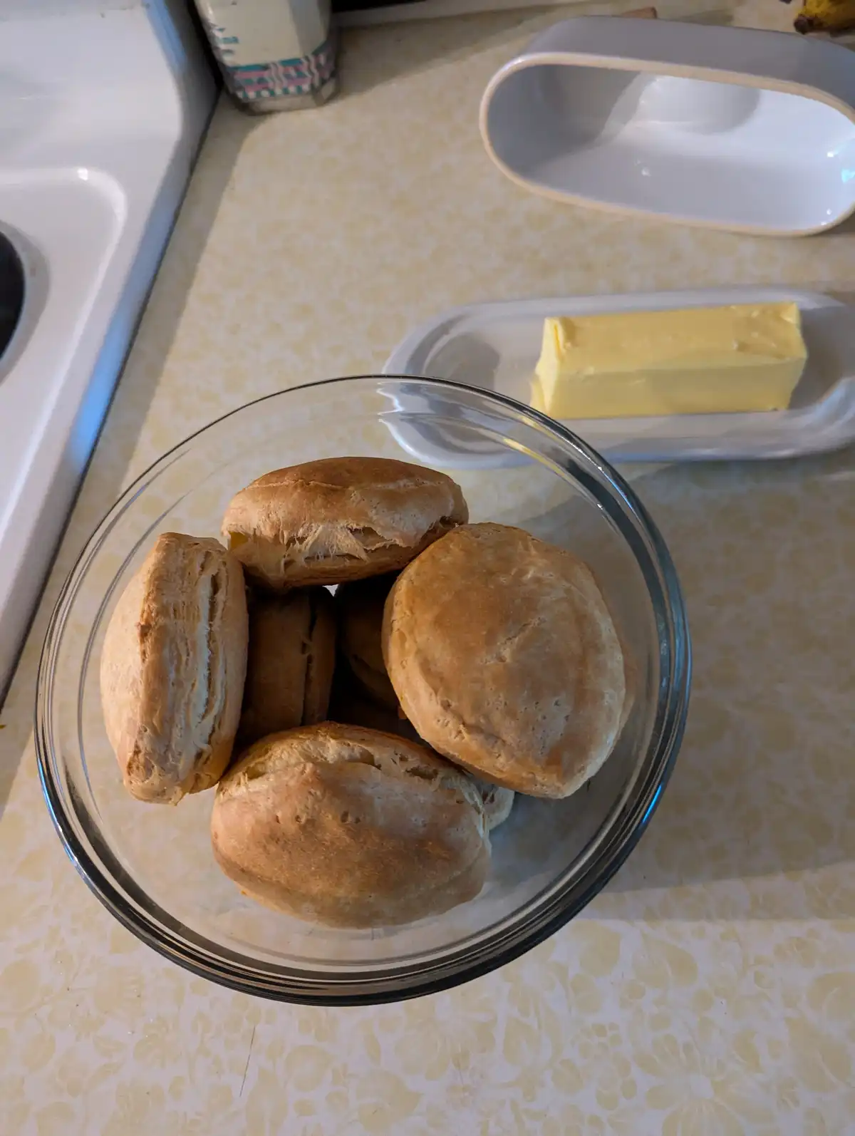 A clear glass bowl filled with golden-brown, freshly baked biscuits sits on a patterned kitchen countertop next to a butter…