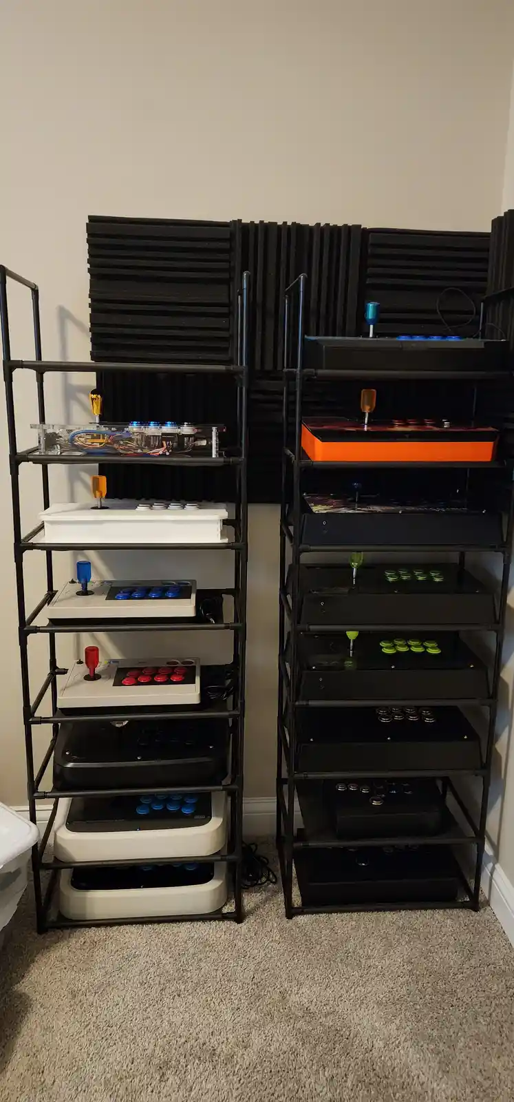 Two tall black metal shelving units are filled with numerous arcade-style game controllers and cabinets, stacked vertically…