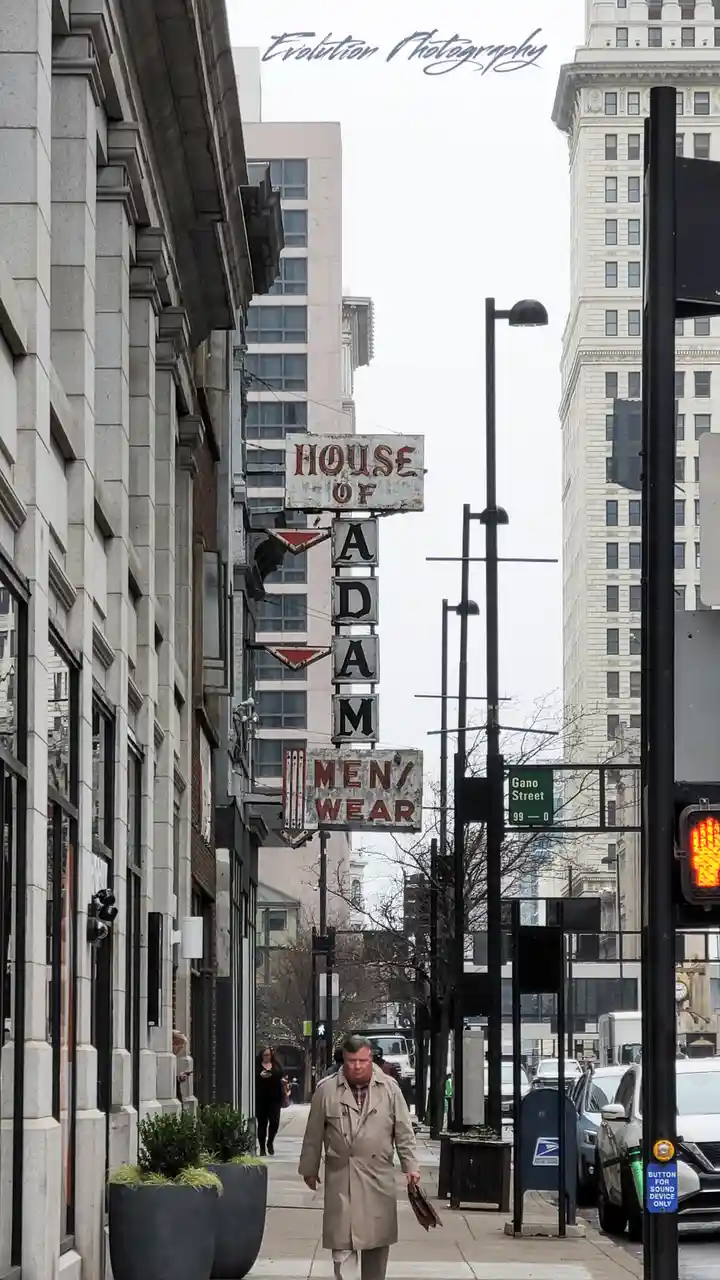 A man in a beige trench coat walks down a city sidewalk past a vintage, weathered vertical sign that reads 'HOUSE OF ADAM…