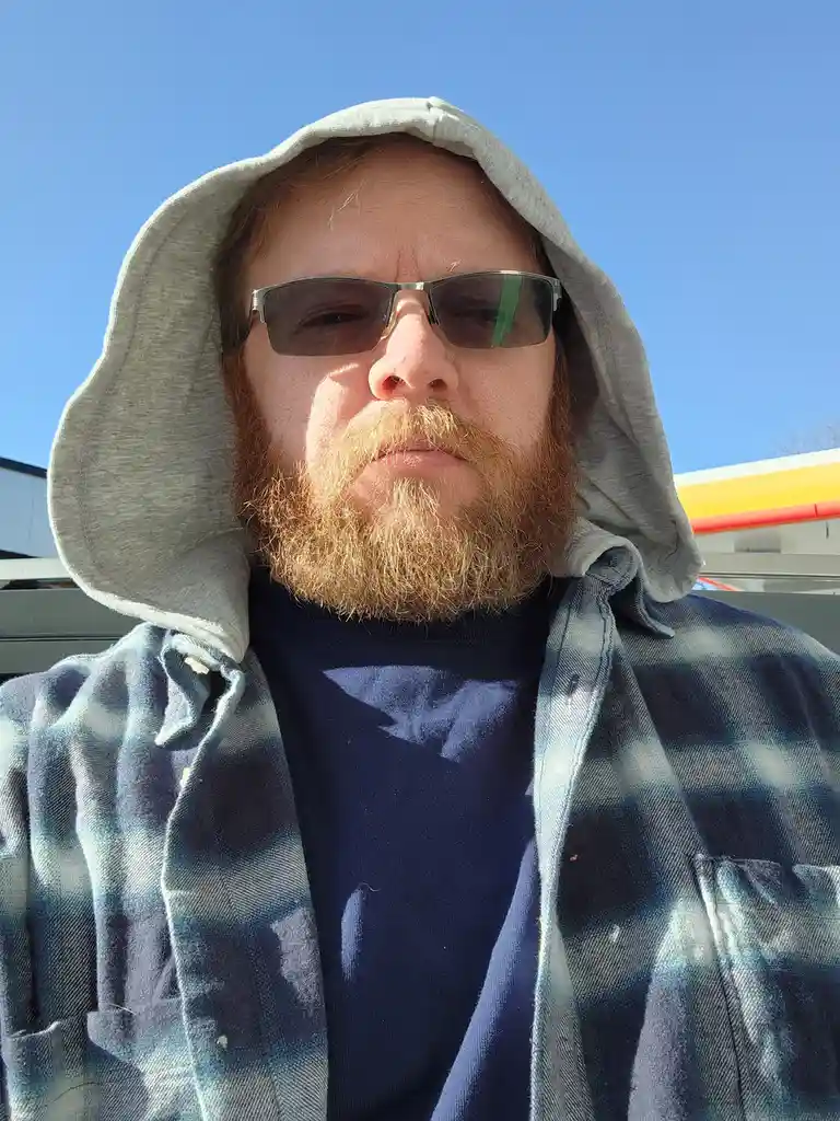 A man with a red beard and sunglasses wears a grey hoodie pulled over his head and a plaid shirt over a t-shirt, standing…