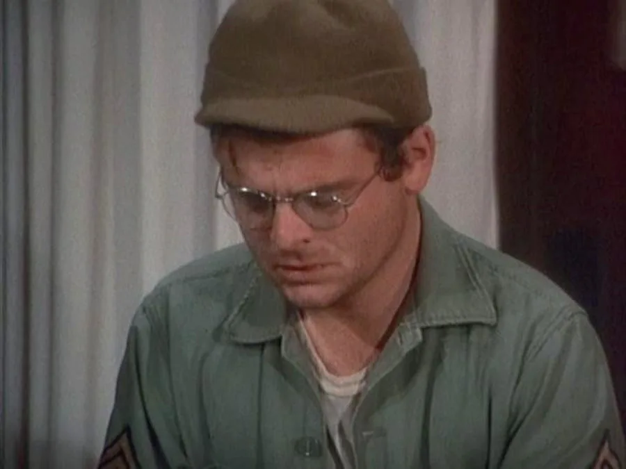 a man in a military-style uniform and cap, wearing glasses, looks down with a visibly frustrated or annoyed expression. The…