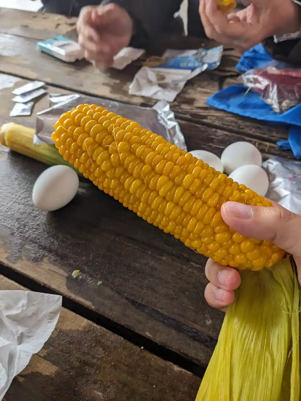 A hand holds a large, glossy yellow corn cob on a wooden table, surrounded by several white eggs. In the background, another…