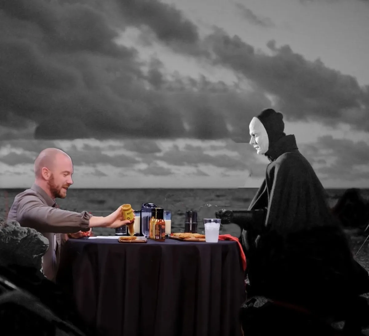 A man in a grey shirt sits at a table by the ocean, smiling as he pours mustard onto his food, while across from him sits a…