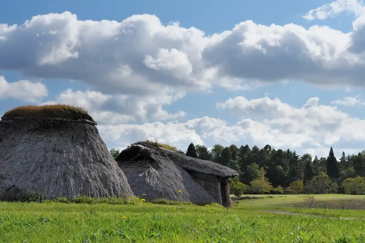 A serene landscape featuring two traditional thatched-roof huts in a grassy field under a partly cloudy blue sky, with trees…