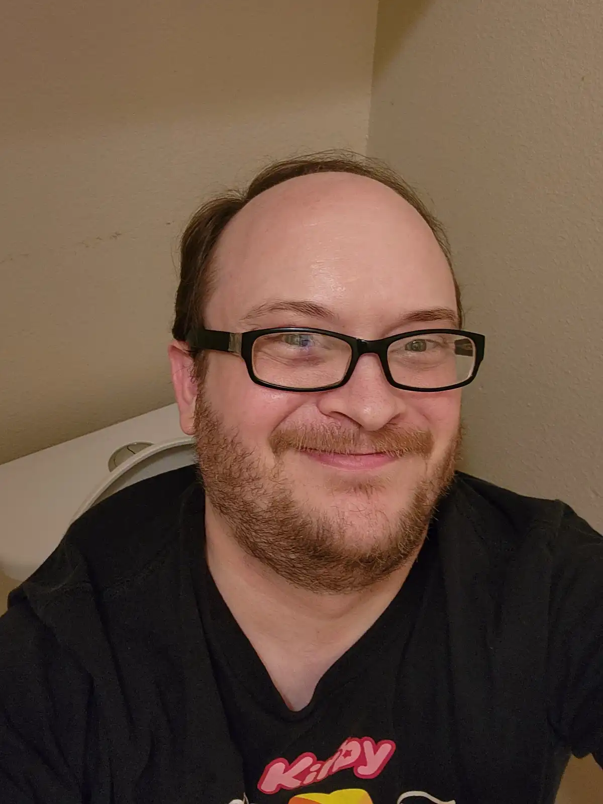 A man with a receding hairline and glasses smiles at the camera while sitting on a toilet, wearing a black t-shirt with a…