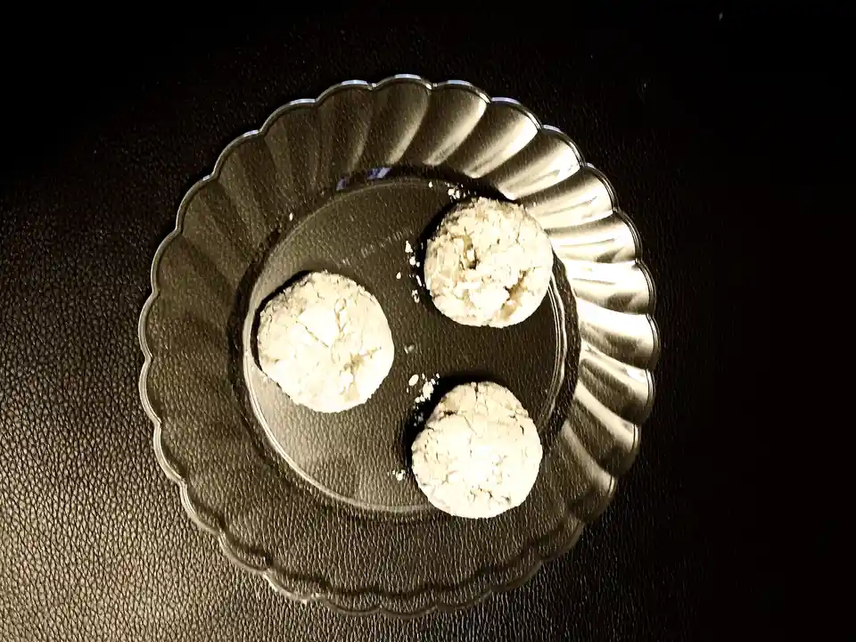 Three round, crumbly, white cookies are arranged on a clear, scalloped glass plate set against a dark, textured surface. The…