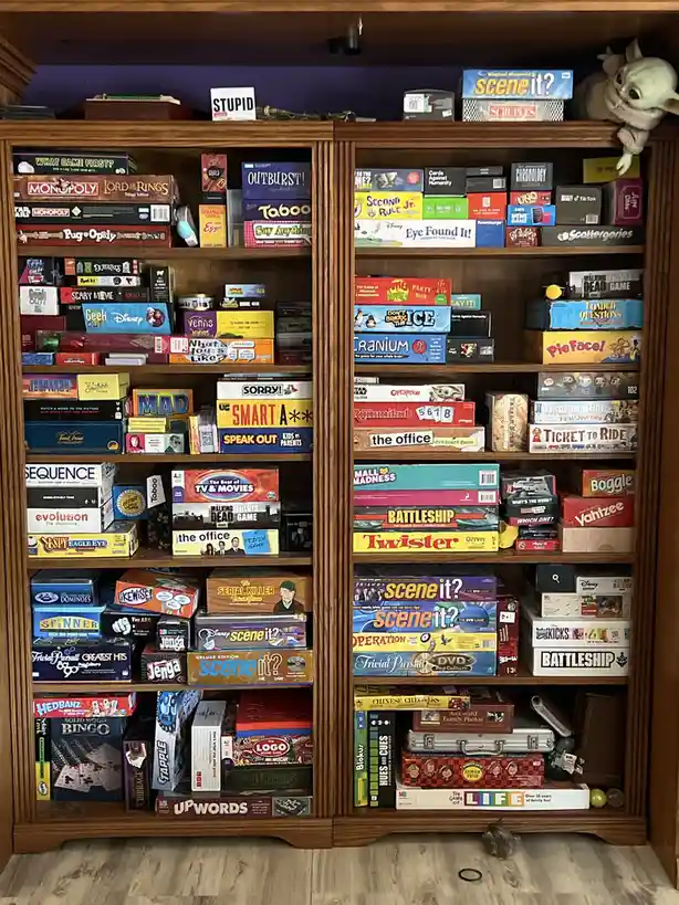 A wooden bookshelf is densely packed with hundreds of colorful board game boxes, including Monopoly, Scrabble, Trivial…