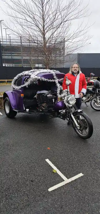 A man in a red and white Santa jacket stands beside a purple, three-wheeled motorcycle that's decorated with silver tinsel…