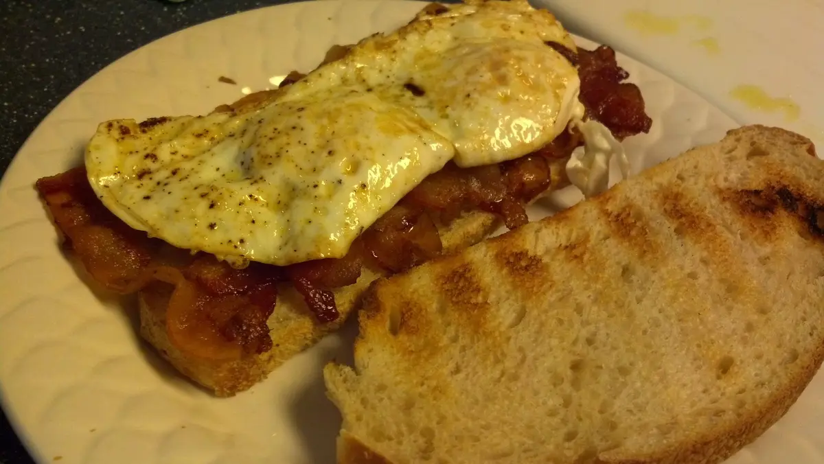 A close-up photo shows a sandwich on a white plate, made with two slices of toasted bread, topped with crispy bacon, a fried…