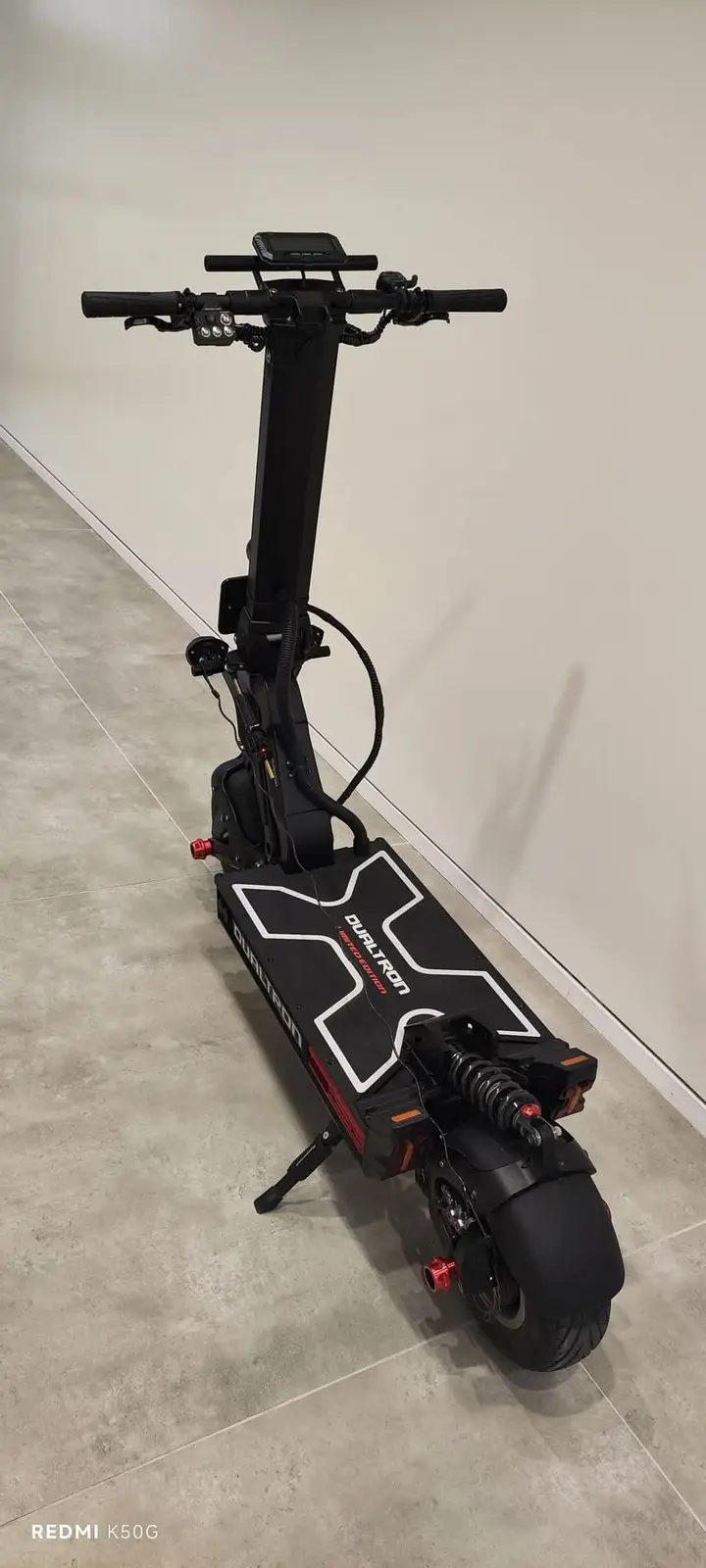 A black Dualtron electric scooter with a large front wheel, suspension, and handlebars is standing upright on a tiled floor…