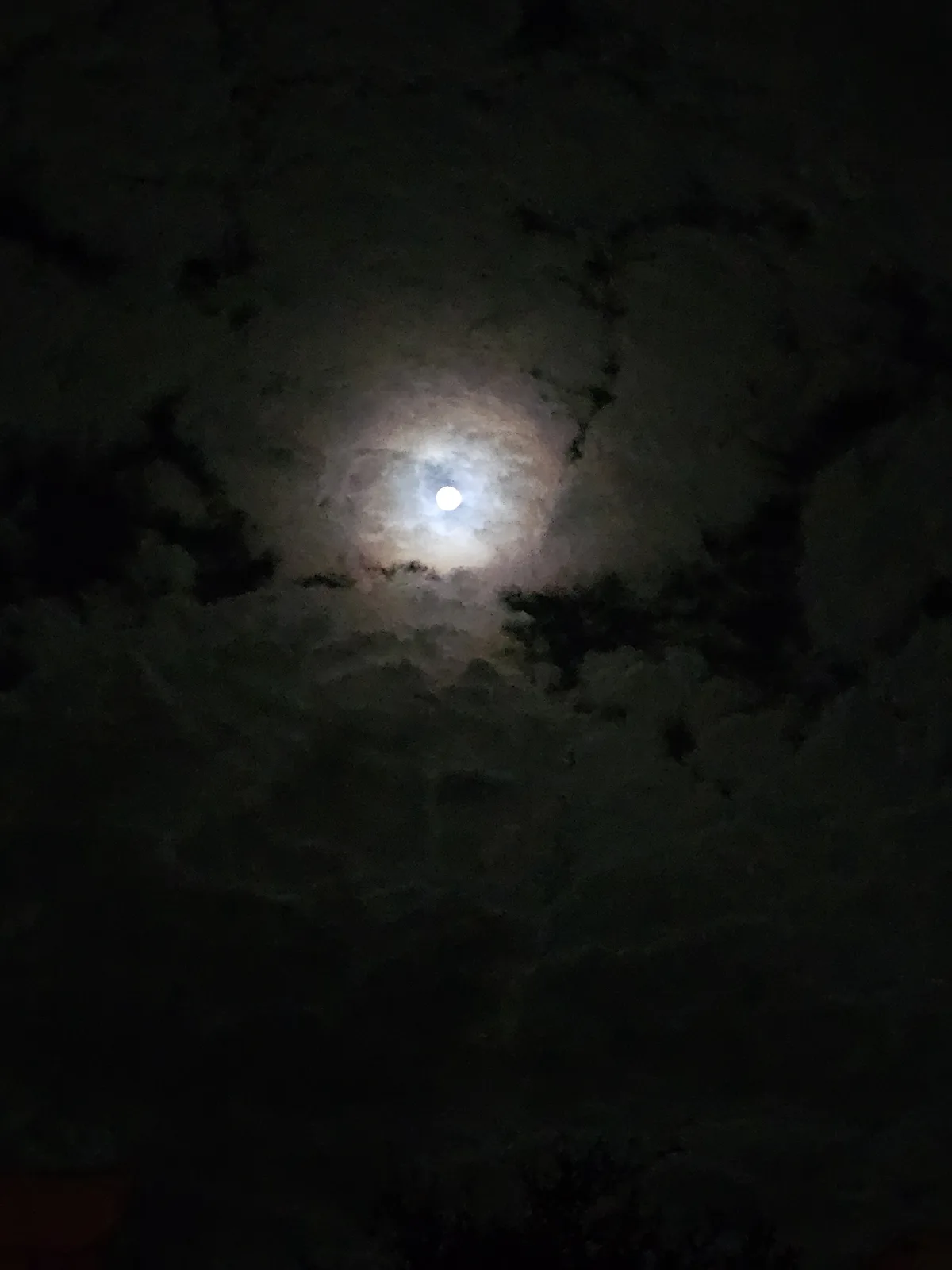 A night sky photo shows a bright, full moon partially obscured by dark, wispy clouds. The moonlight illuminates the clouds…