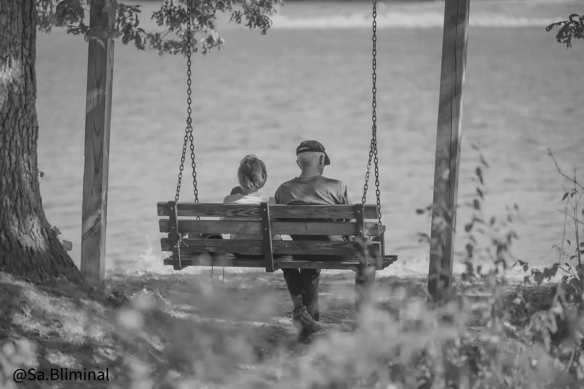 A black-and-white photo shows an older couple sitting on a wooden swing facing away from the camera, overlooking a body of…