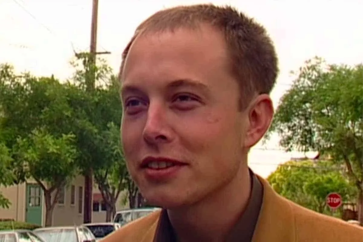 A close-up shot of a young man with short brown hair and a slight smile, wearing a tan jacket over a dark shirt, standing…