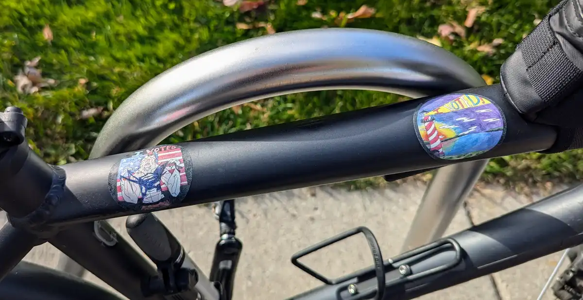 A close-up of a black bicycle handlebar with two colorful 'I VOTED' stickers. One sticker features a cartoon Santa Claus…
