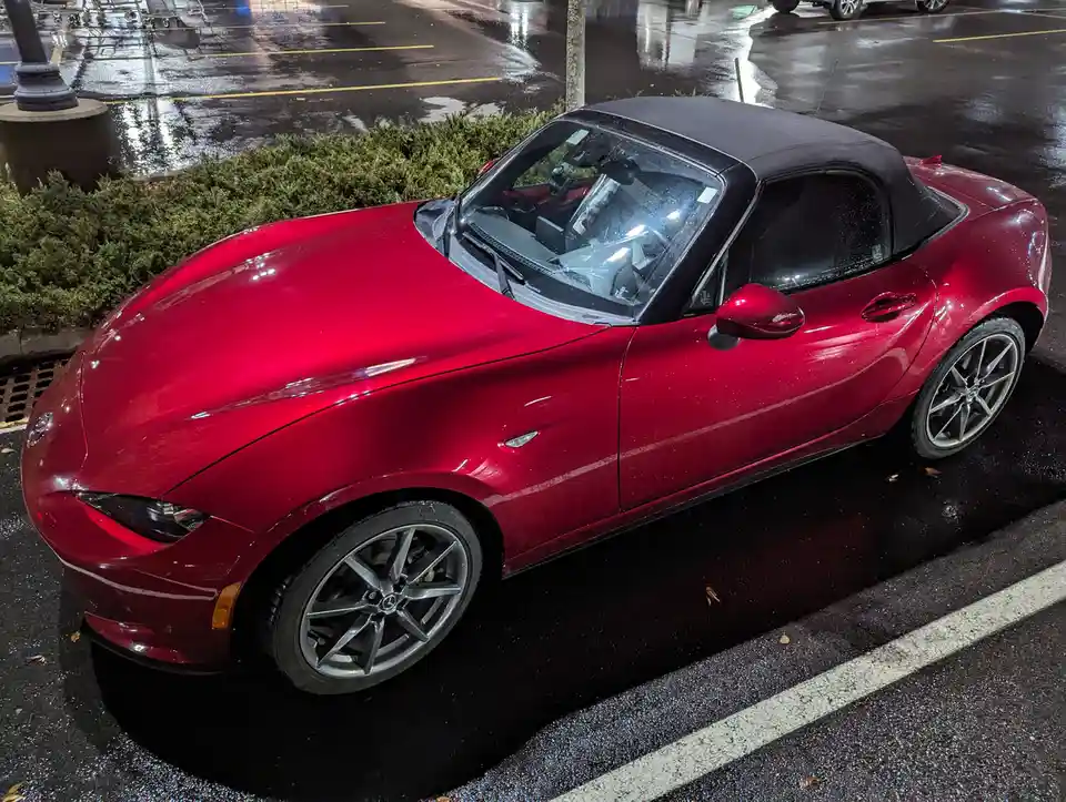 A red Mazda Miata convertible is parked in a wet parking lot at night, its black soft top closed. The car's glossy paint…