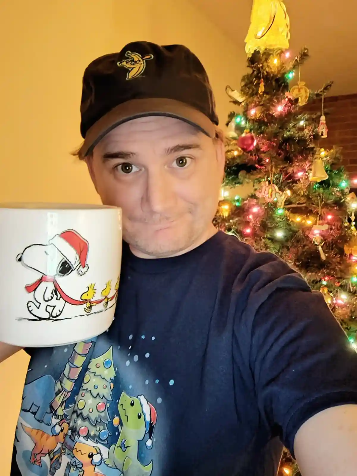 A man in a dark cap and festive t-shirt holds a white mug featuring Snoopy in a Santa hat, standing in front of a lit…