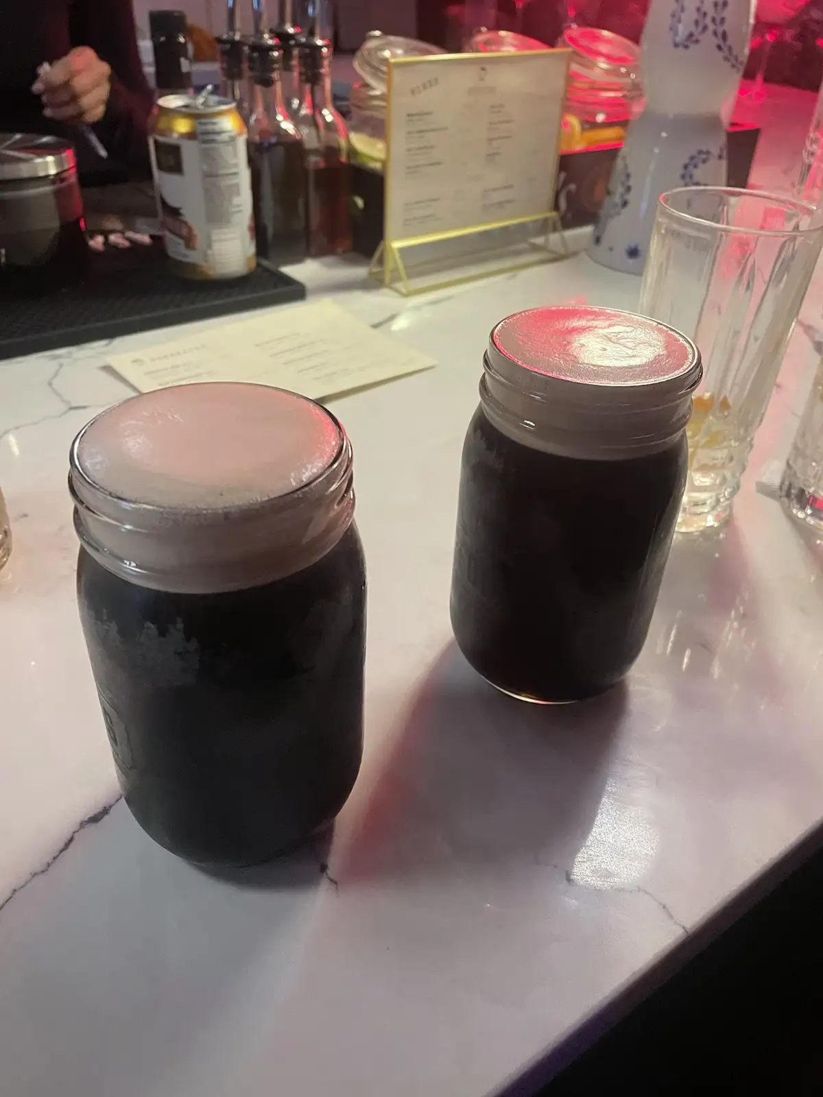 Two mason jars filled with dark liquid and topped with a thick, creamy head sit on a white marble bar counter. The…