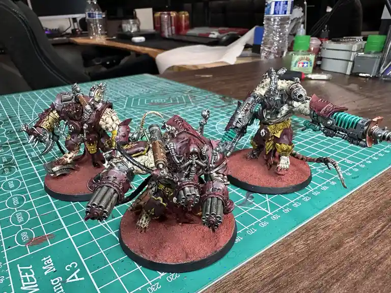 Three heavily armed, custom-painted miniature figures resembling grotesque, biomechanical rat-like warriors stand on red…