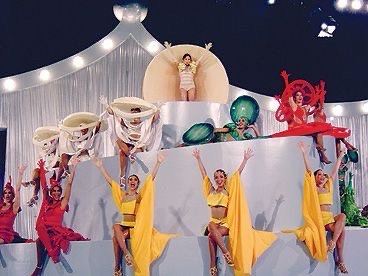 A group of performers in yellow and red costumes strike dramatic poses on a tiered stage with large, stylized props…