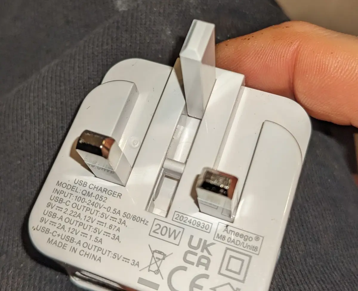A close-up photo shows a white UK-style USB wall charger held in a person's hand, with printed technical specifications…
