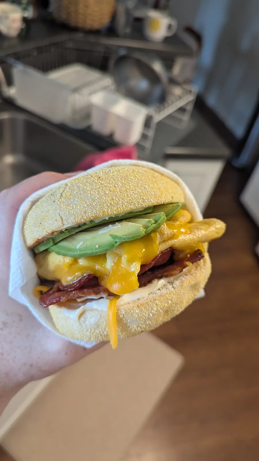 A hand holds a generously filled breakfast sandwich with a toasted English muffin, melted yellow cheese, crispy bacon, a…