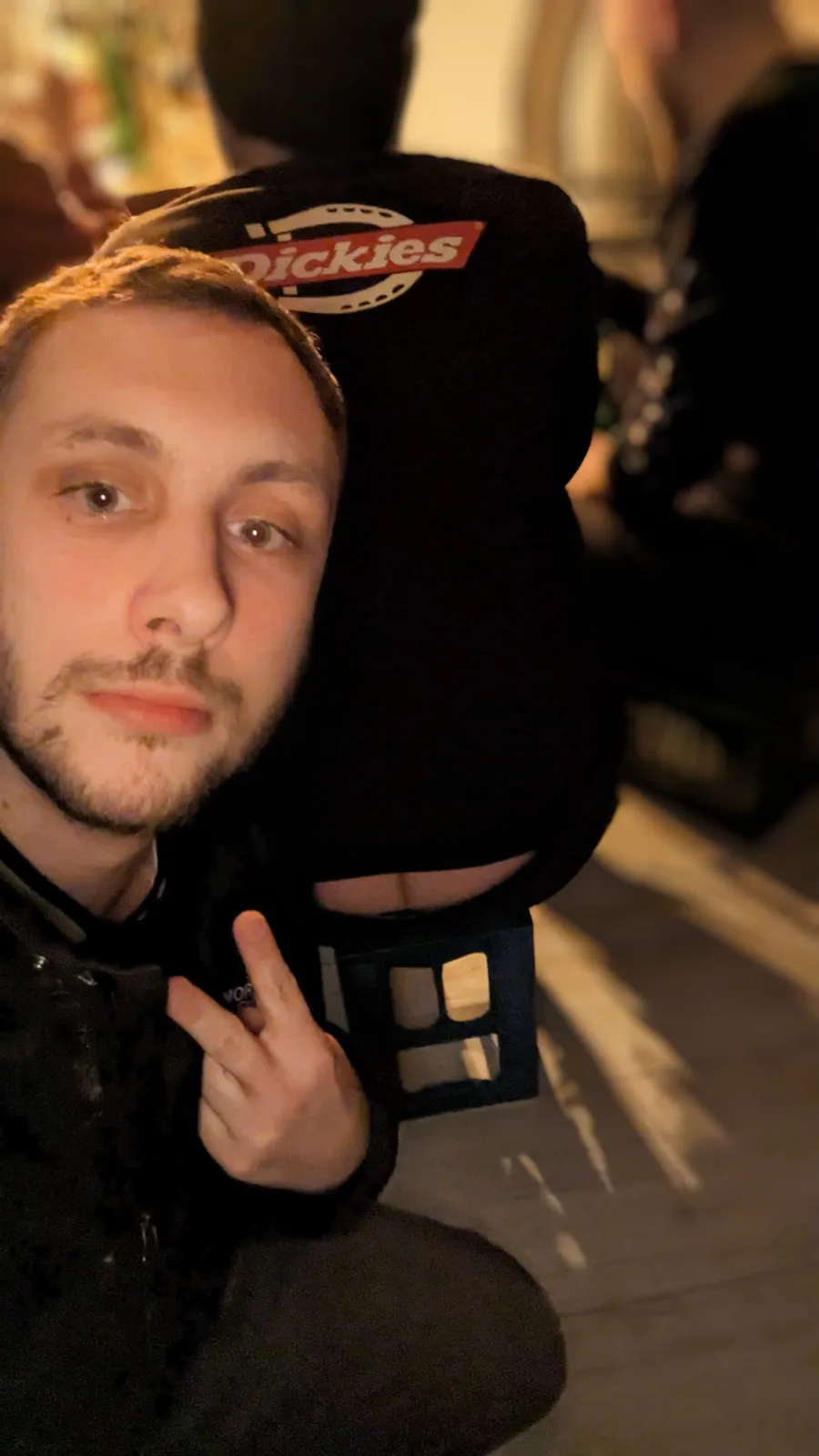 A man with a beard and short hair makes a peace sign while looking at the camera, wearing a black jacket with a 'Dickies'…