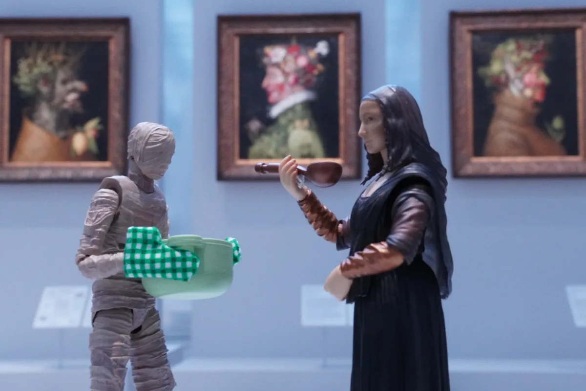 Two action figures stand in a museum gallery with framed paintings on the wall behind them. One figure is wrapped in grey…
