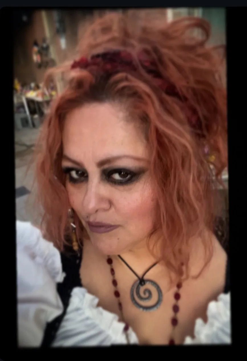 A woman with reddish hair, dark eye makeup, and a spiral pendant necklace stares intensely at the camera. She wears a white…