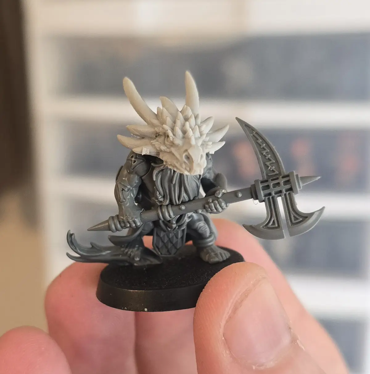 A close-up photo shows a person holding a small, unpainted fantasy miniature of a humanoid figure with a dragon skull for a…