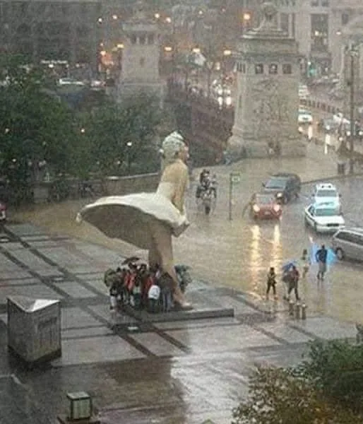A large statue of a woman in a flowing white dress stands in a rainy city square, her skirt billowing outward as if caught…