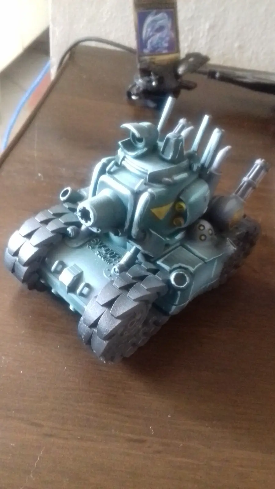 A heavily modified, teal-colored toy tank with multiple cannons and spikes sits on a wooden table. The overlaid text reads…