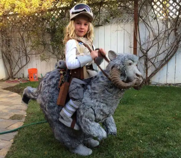 A young girl in a Star Wars pilot costume, complete with goggles and utility belt, is posing while riding a large, furry…