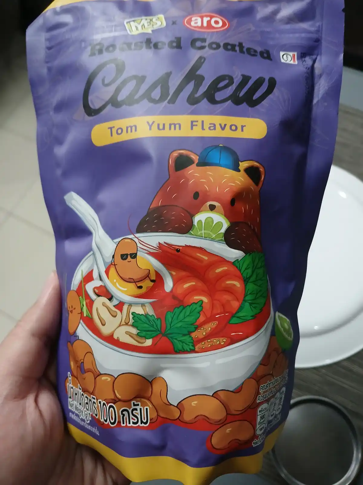 A hand holds a purple bag of 'YES x aro' Roasted Coated Cashew in Tom Yum Flavor. The packaging features cartoon…