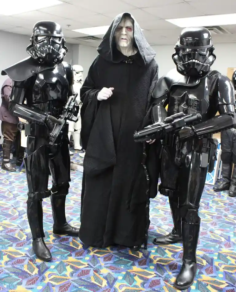Three people in Star Wars costumes pose indoors: a central figure in a black hooded robe with a pale, scarred face (Darth…