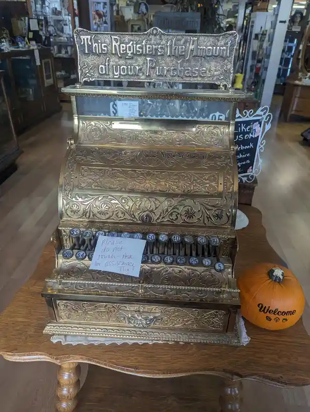 An ornate, vintage brass cash register with a sign reading 'This Registers the Amount of your Purchase' sits on a wooden…