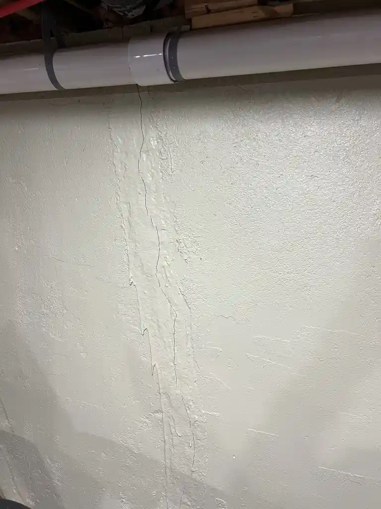 A close-up photo shows a vertical crack running down a textured white wall, with a white PVC pipe running horizontally…