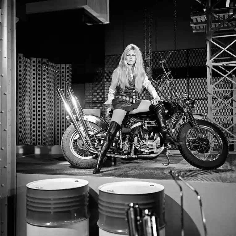 A black-and-white photograph shows a blonde woman with long hair sitting on a large, chrome motorcycle in an…