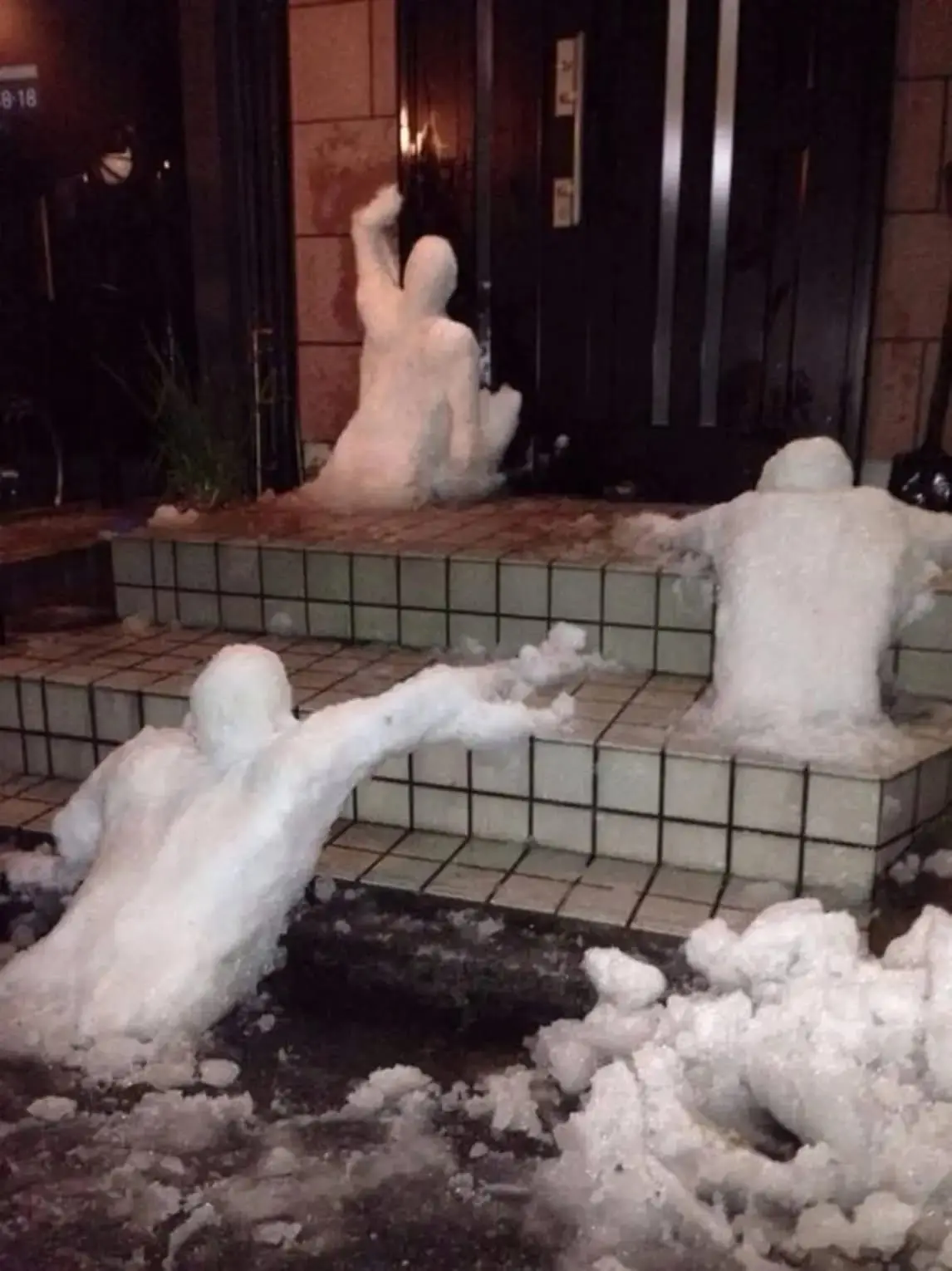 Three snowmen are arranged on tiled steps outside a building at night, appearing to be in a chaotic fight. One snowman is on…