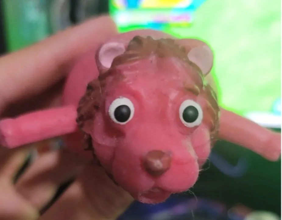 A close-up photo shows a person's hand holding a pink plastic toy animal with brown spiky hair, large white-rimmed eyes, and…