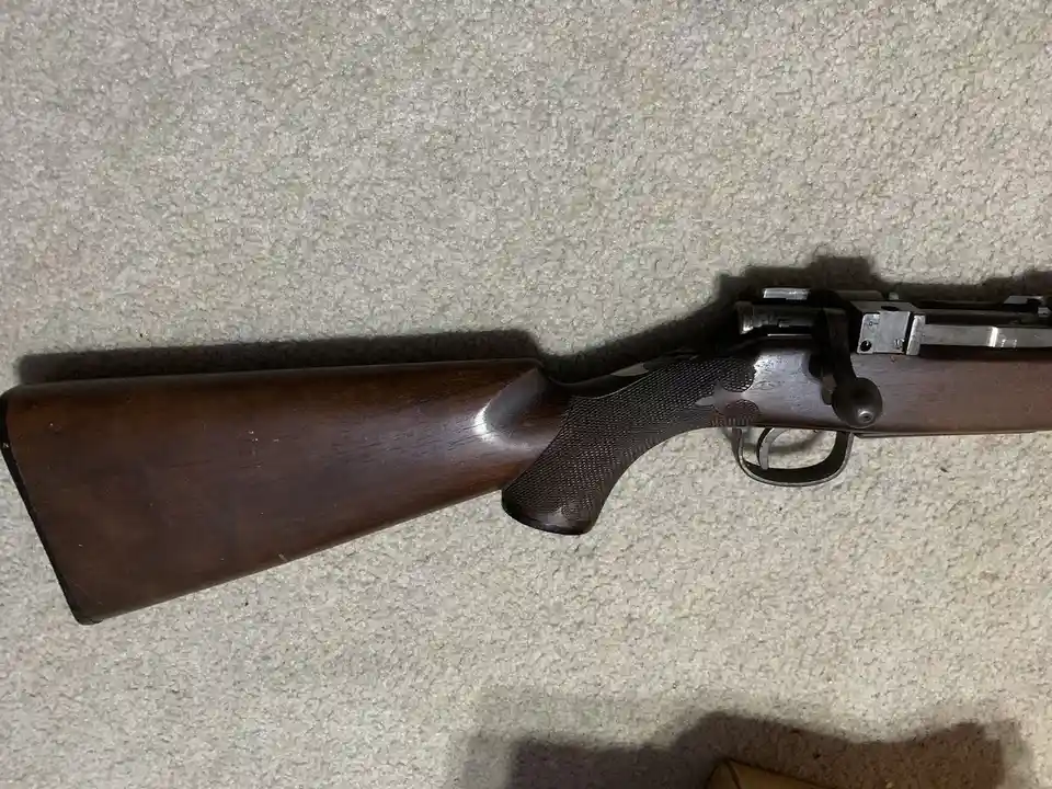 A close-up photo of a wooden-stocked rifle lying horizontally on a light-colored carpet, showing its trigger, grip, and…