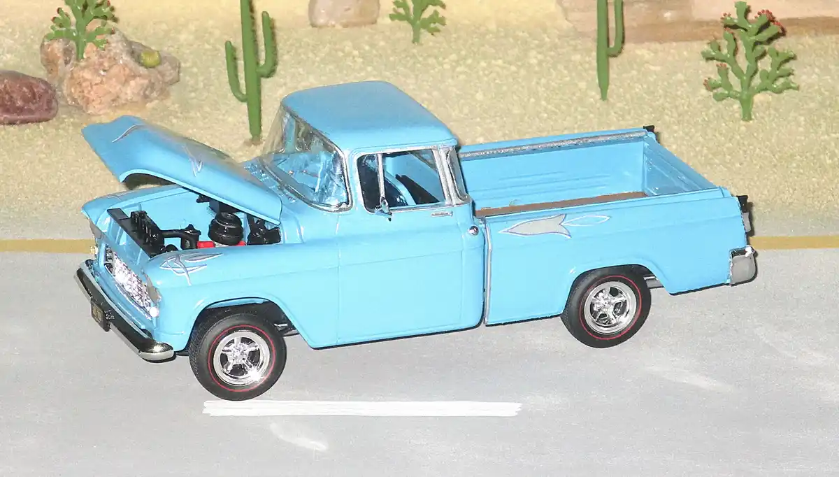 A light blue vintage toy pickup truck with its hood open, parked on a grey surface with a desert diorama background…