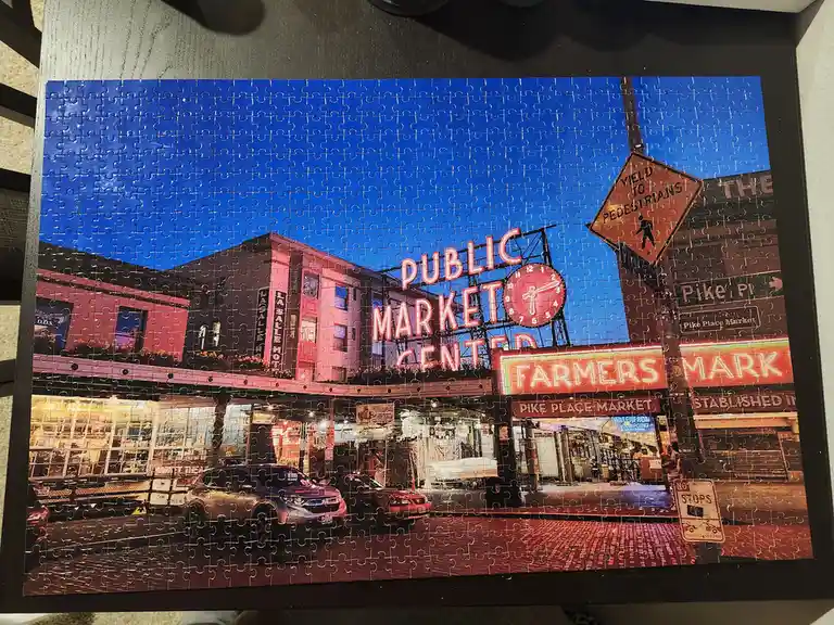 A completed jigsaw puzzle depicting the iconic Pike Place Market in Seattle, showing its famous neon sign, a large clock…