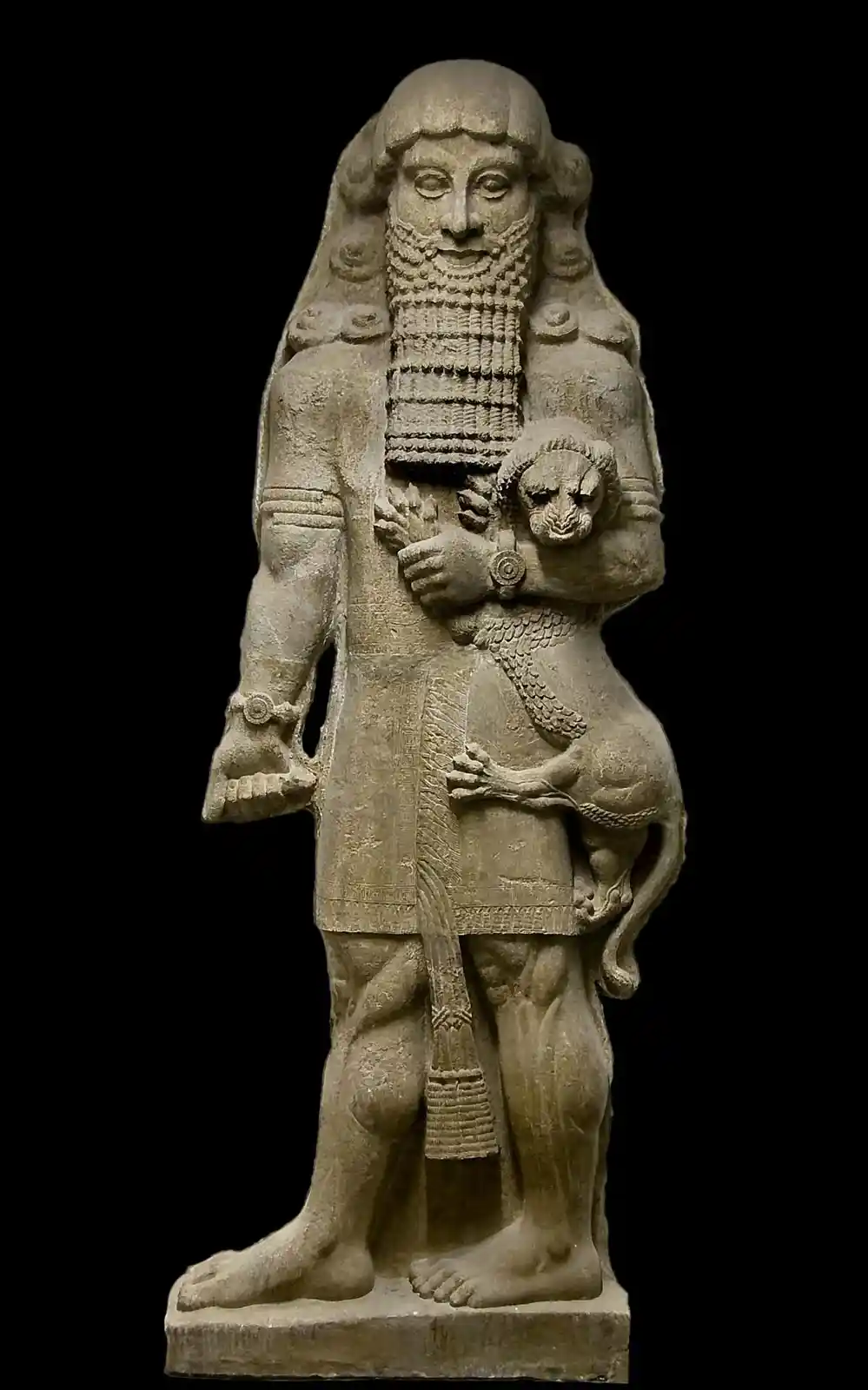 A stone statue of a bearded, robed ancient figure holding a small lion cub in his arms, set against a black background. The…