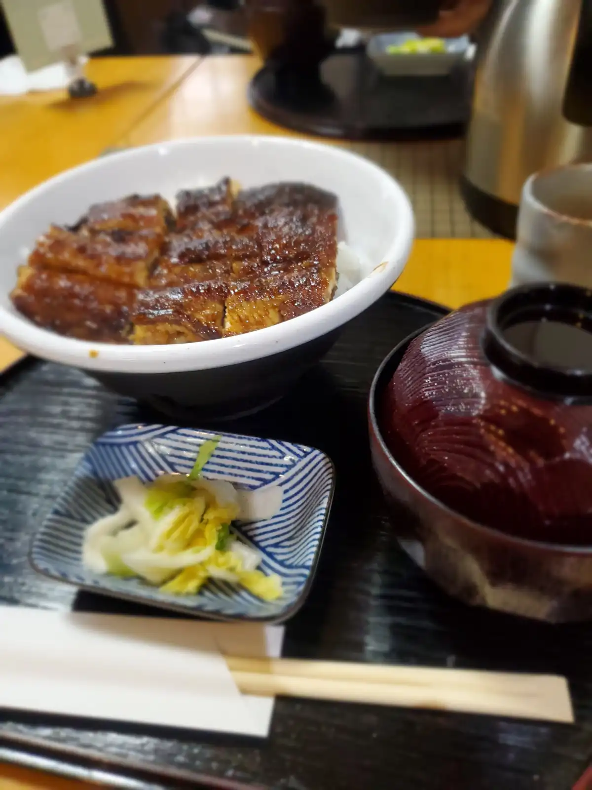 A close-up photo of a Japanese meal featuring a bowl of unagi donburi with grilled eel over rice, accompanied by a small…