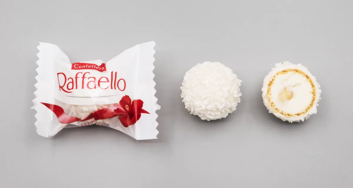 A Raffaello candy wrapper sits next to a whole coconut-covered candy and a half-eaten one revealing its creamy filling, all…